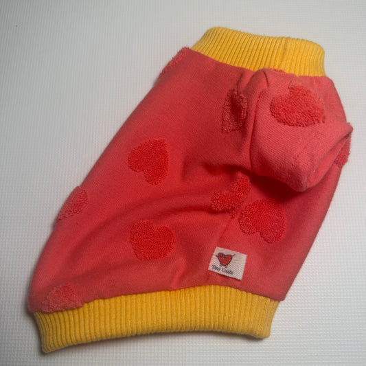 A small pink pet sweater with red heart details and yellow ribbed trim, laid flat on a light gray textured background.